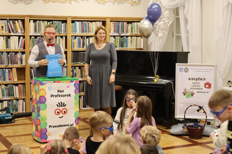 kolorowe zdjęcie, 2 osoby dorosłe, napis Noc Bibliotek, Pan Profesorek, dzieci siedzą na dywanie i poduszkach,