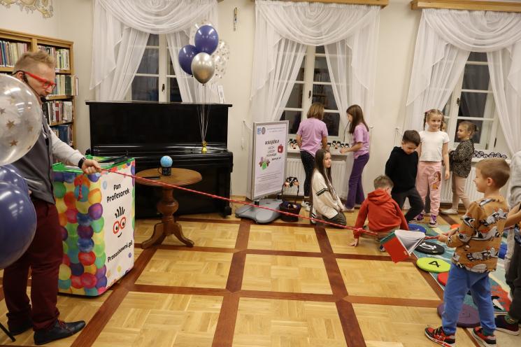 kolorowe zdjęcie, 2 osoby dorosłe, napis Noc Bibliotek, Pan Profesorek, dzieci siedzą na dywanie i poduszkach,