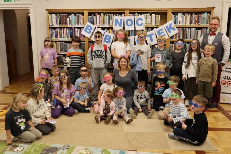 kolorowe zdjęcie, 2 osoby dorosłe, napis Noc Bibliotek, Pan Profesorek, dzieci siedzą na dywanie i poduszkach,