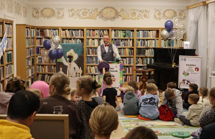 kolorowe zdjęcie, 2 osoby dorosłe, napis Noc Bibliotek, Pan Profesorek, dzieci siedzą na dywanie i poduszkach,