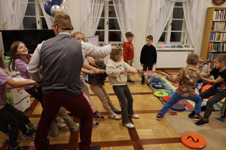 kolorowe zdjęcie, 2 osoby dorosłe, napis Noc Bibliotek, Pan Profesorek, dzieci siedzą na dywanie i poduszkach,