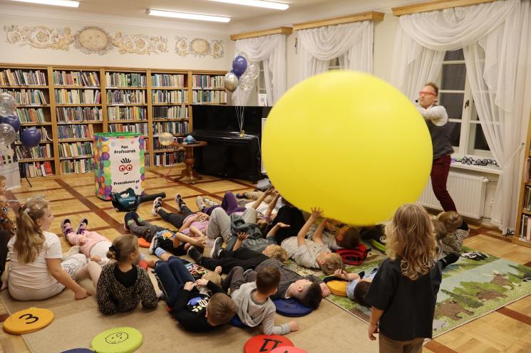 kolorowe zdjęcie, 2 osoby dorosłe, napis Noc Bibliotek, Pan Profesorek, dzieci siedzą na dywanie i poduszkach,