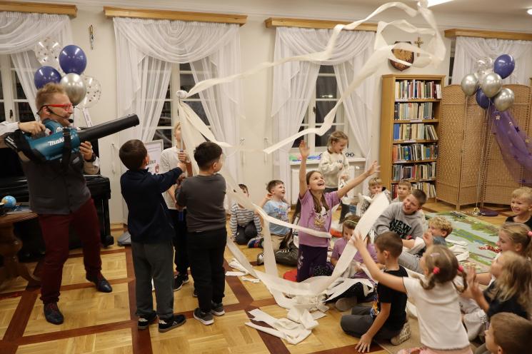 kolorowe zdjęcie, 2 osoby dorosłe, napis Noc Bibliotek, Pan Profesorek, dzieci siedzą na dywanie i poduszkach,