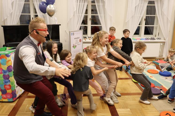 kolorowe zdjęcie, 2 osoby dorosłe, napis Noc Bibliotek, Pan Profesorek, dzieci siedzą na dywanie i poduszkach,