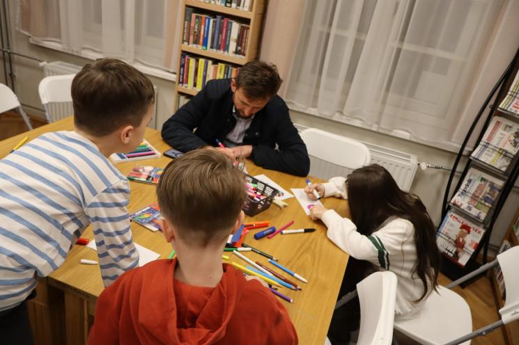 kolorowe zdjęcie, 2 osoby dorosłe, napis Noc Bibliotek, Pan Profesorek, dzieci siedzą na dywanie i poduszkach,