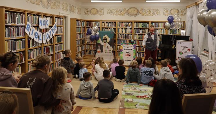 kolorowe zdjęcie, 2 osoby dorosłe, napis Noc Bibliotek, Pan Profesorek, dzieci siedzą na dywanie i poduszkach,