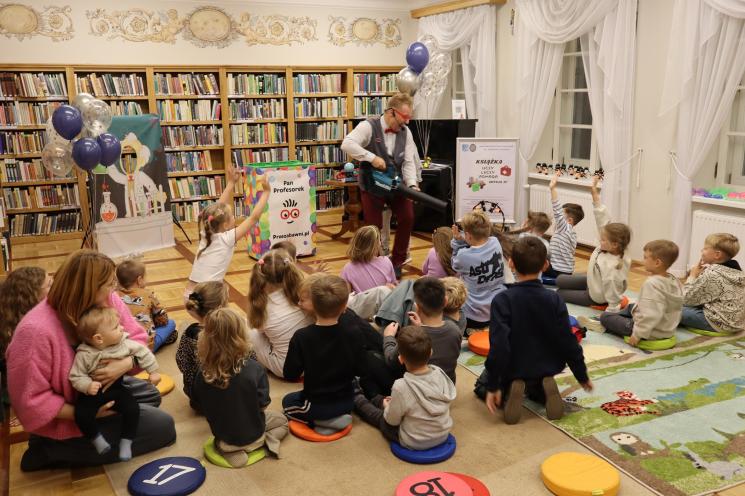 kolorowe zdjęcie, 2 osoby dorosłe, napis Noc Bibliotek, Pan Profesorek, dzieci siedzą na dywanie i poduszkach,