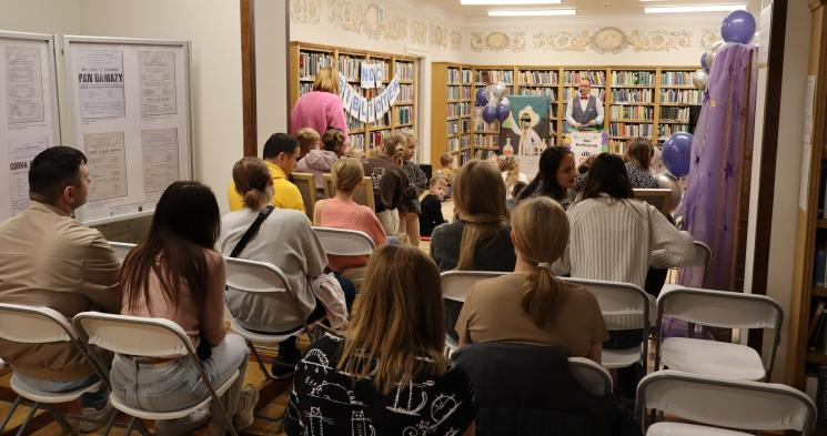 kolorowe zdjęcie, 2 osoby dorosłe, napis Noc Bibliotek, Pan Profesorek, dzieci siedzą na dywanie i poduszkach,