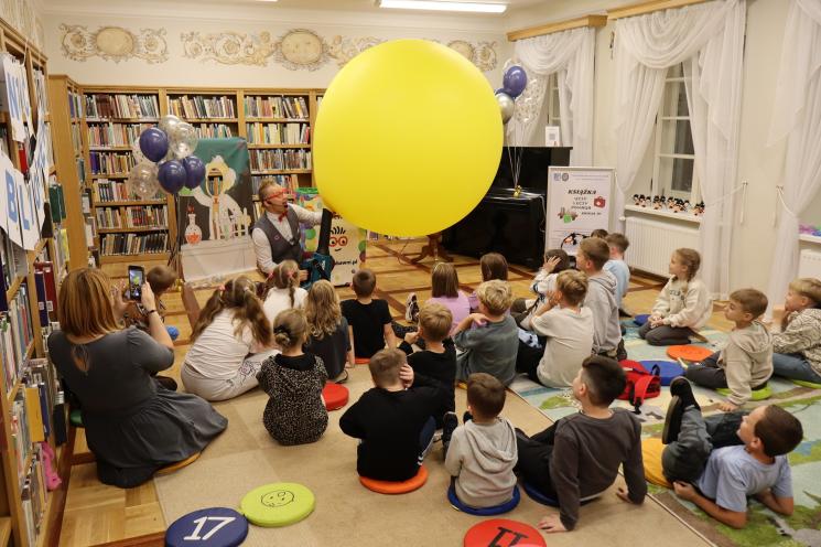 kolorowe zdjęcie, 2 osoby dorosłe, napis Noc Bibliotek, Pan Profesorek, dzieci siedzą na dywanie i poduszkach,