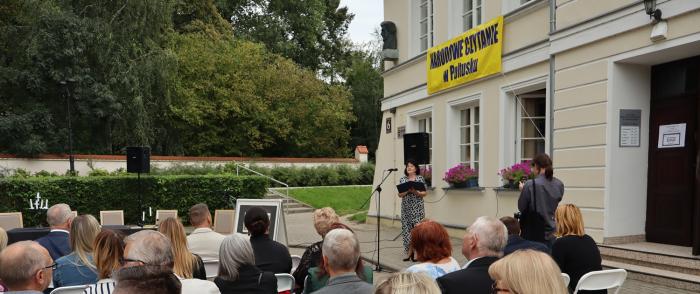 Grupa ludzi siedzi na krzesłach w plenerze, na tle budynku biblioteki, na budynku napis Narodowe Czytanie. 