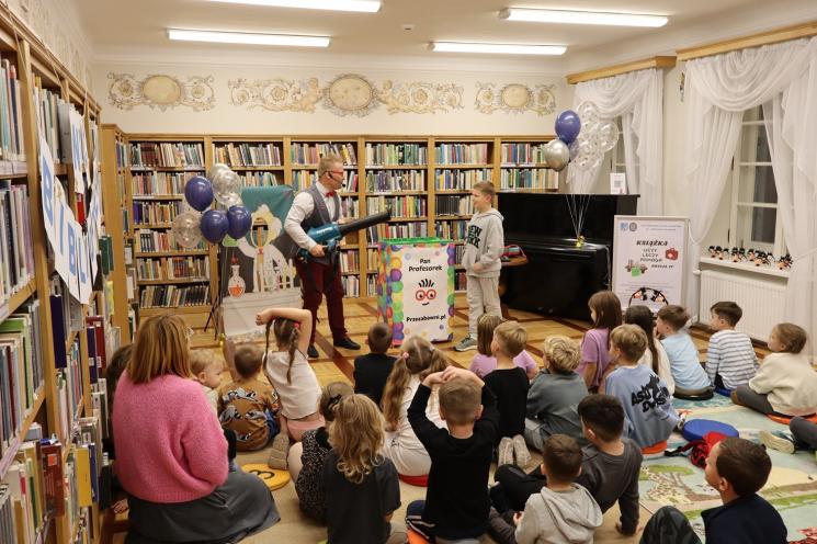 kolorowe zdjęcie, 2 osoby dorosłe, napis Noc Bibliotek, Pan Profesorek, dzieci siedzą na dywanie i poduszkach,