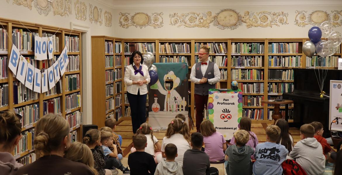kolorowe zdjęcie, 2 osoby dorosłe, napis Noc Bibliotek, Pan Profesorek, dzieci siedzą na dywanie i poduszkach,