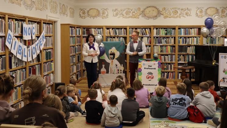kolorowe zdjęcie, 2 osoby dorosłe, napis Noc Bibliotek, Pan Profesorek, dzieci siedzą na dywanie i poduszkach,