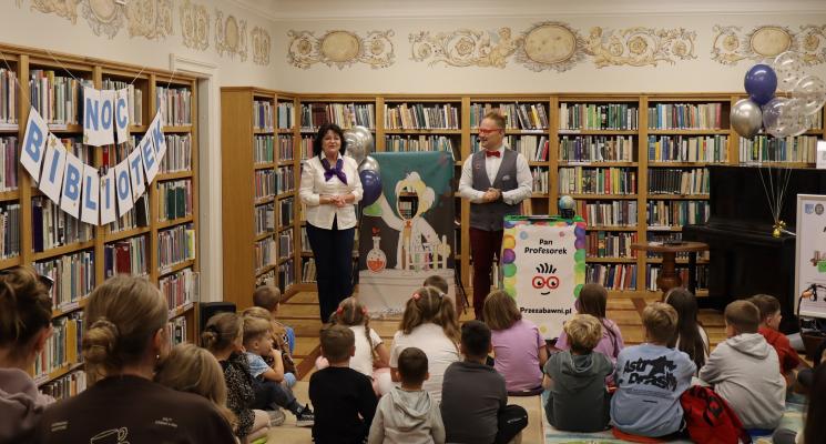 kolorowe zdjęcie, 2 osoby dorosłe, napis Noc Bibliotek, Pan Profesorek, dzieci siedzą na dywanie i poduszkach,