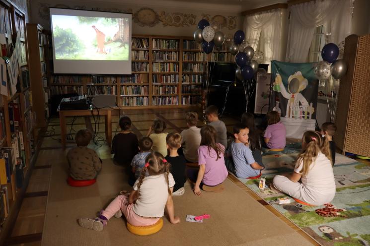 kolorowe zdjęcie, 2 osoby dorosłe, napis Noc Bibliotek, Pan Profesorek, dzieci siedzą na dywanie i poduszkach,